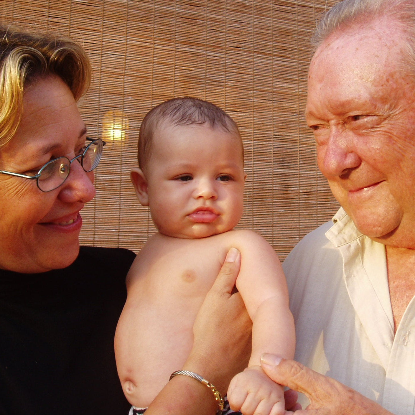 Woman holding a baby with an older man standing next to them, all smiling outdoors.